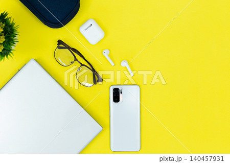 Bright yellow background holds tech and style. White phone rests near wireless earbuds and glasses. Laptop corner shows active workspace use. Bitcoin coin adds modern finance or digital theme 140457931