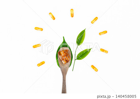 Golden capsules rest on a wooden spoon. Green leaves frame the natural supplement display. White background emphasizes clarity and purity. The image suggests health, wellness, and nature's power 140458005