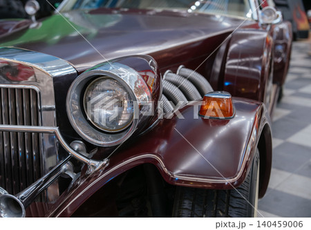 Detailed close-up of vintage maroon car showcasing chrome accents and a prominent headlight. Reflective surfaces highlight intricate design elements, while the blurred background suggests an indoor 140459006