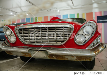 Close-up view of classic red car showcasing its chrome grille and dual headlights. Background features colorful wall art, enhancing the vintage automobile's charm in an indoor setting. 140459012