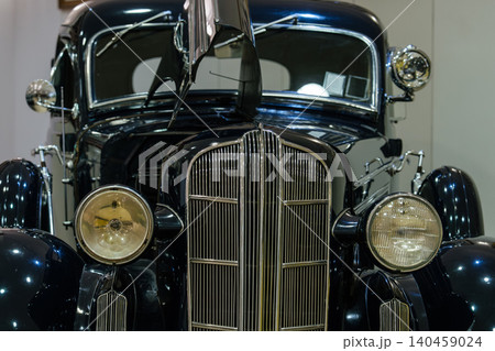 Detailed front view of classic dark blue automobile showcasing chrome grille, circular headlights, and partially open hood. Indoor setting highlights the car's glossy finish and elegant design. Detailed front view of classic dark blue automobile showcasing chrome grille, circular headlights, and partially open hood. Indoor setting highlights the car's glossy finish and elegant design. 140459024
