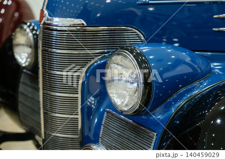 Detailed close-up showcases front right side of vintage blue automobile. Focus on round headlight with chrome rim and multi-slat chrome grille, reflecting studio lights. 140459029