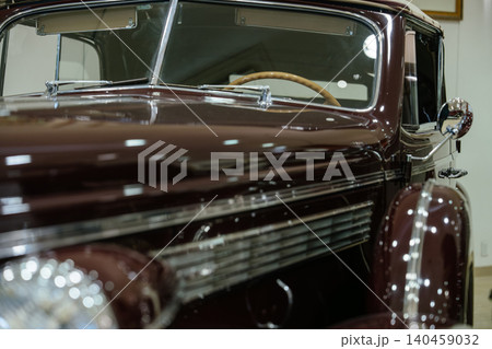 Close-up shot captures vintage maroon automobile showcasing shiny paint and chrome details. Interior features wooden steering wheel and light-colored fabric, suggesting luxury in a museum setting. Close-up shot captures vintage maroon automobile showcasing shiny paint and chrome details. Interior features wooden steering wheel and light-colored fabric, suggesting luxury in a museum setting. 140459032