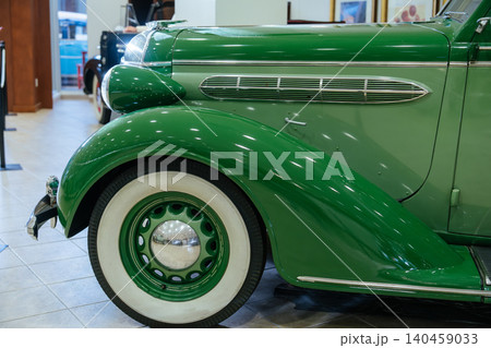 Detailed view of front fender and side panel of shiny green vintage car. Features whitewall tires, chrome hubcap, and intricate chrome detailing, set in a museum or exhibition space. Detailed view of front fender and side panel of shiny green vintage car. Features whitewall tires, chrome hubcap, and intricate chrome detailing, set in a museum or exhibition space. 140459033