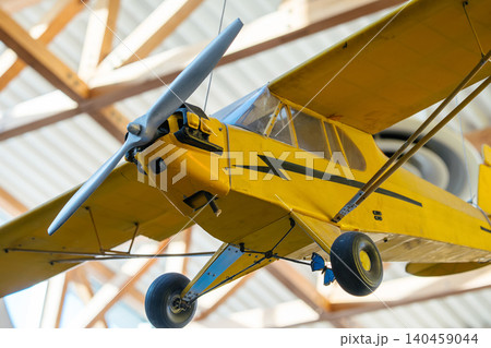 Close-up view of a vintage yellow small airplane hanging from the ceiling indoors. Features include a silver propeller, transparent cockpit, and black markings, with sunlight illuminating the scene. 140459044