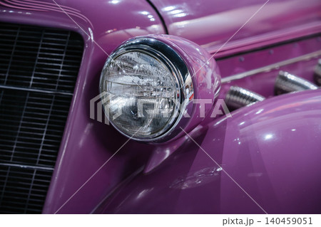 Detailed close-up captures vintage purple automobile's round headlight with chrome rim and textured lens beside a black horizontal-bar grille. Bright lighting enhances glossy finish and chrome exhaust Detailed close-up captures vintage purple automobile's round headlight with chrome rim and textured lens beside a black horizontal-bar grille. Bright lighting enhances glossy finish and chrome exhaust 140459051