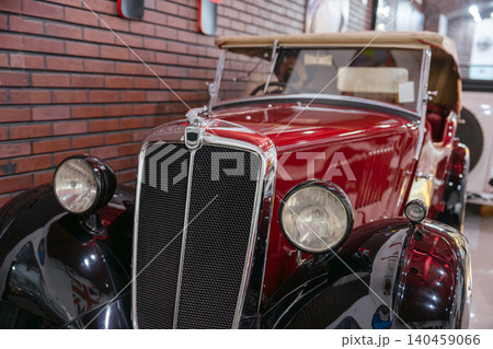 Close-up view highlights front of vintage red convertible car with chrome grille and round headlights. Glossy red body and tan fabric top stand out against textured brick wall backdrop. Close-up view highlights front of vintage red convertible car with chrome grille and round headlights. Glossy red body and tan fabric top stand out against textured brick wall backdrop. 140459066