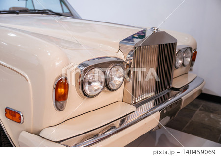 Frontal close-up of antique cream-colored luxury car showcasing iconic chrome grille and dual round headlights. Bright lighting highlights polished surfaces against a clean background. Frontal close-up of antique cream-colored luxury car showcasing iconic chrome grille and dual round headlights. Bright lighting highlights polished surfaces against a clean background. 140459069