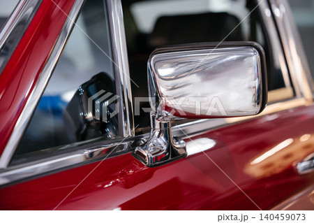 Detailed close-up view of shiny chrome side mirror attached to door of classic red automobile. Polished surface reflects light and distorted images of surroundings, showcasing vintage design. Detailed close-up view of shiny chrome side mirror attached to door of classic red automobile. Polished surface reflects light and distorted images of surroundings, showcasing vintage design. 140459073