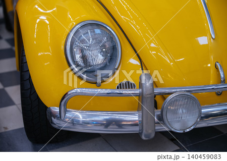 Closeup photo of a yellow vintage car with chrome bumper and classic round headlight details. Bright retro automobile with iconic curved design and polished metal reflections inside a car museum. Closeup photo of a yellow vintage car with chrome bumper and classic round headlight details. Bright retro automobile with iconic curved design and polished metal reflections inside a car museum. 140459083