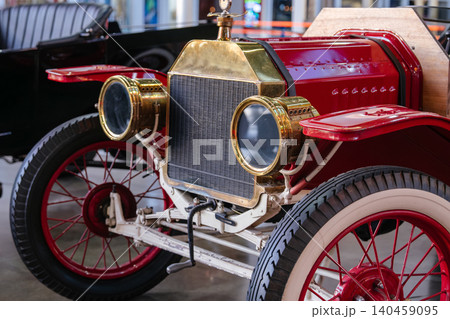 Red antique automobile with brass headlights and vintage spoked wheels displayed inside a classic car museum. Historic retro vehicle features early automotive engineering and elegant transportation Red antique automobile with brass headlights and vintage spoked wheels displayed inside a classic car museum. Historic retro vehicle features early automotive engineering and elegant transportation 140459095