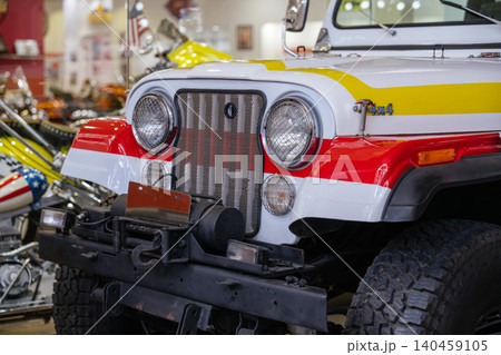 Close-up view of vintage white off-road vehicle featuring bold red and yellow racing stripes, chrome grille, and round headlights. Partially visible is a custom motorcycle with a star-spangled fender Close-up view of vintage white off-road vehicle featuring bold red and yellow racing stripes, chrome grille, and round headlights. Partially visible is a custom motorcycle with a star-spangled fender 140459105