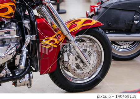 Close-up shot features custom motorcycle's front wheel and fender, showcasing vibrant red color with yellow and orange flame graphics. Polished chrome details enhance the striking design, with another 140459119