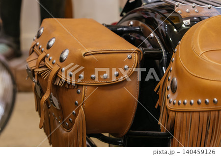 Close-up of a brown leather motorcycle saddlebag featuring decorative metal studs and fringes. A portion of a matching motorcycle seat is visible, showcasing craftsmanship and vintage style. 140459126