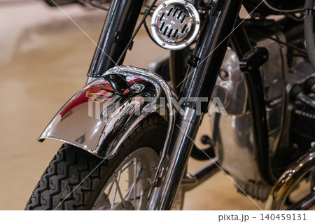 Detailed close-up view of vintage motorcycle front end, showcasing shiny chrome fender, black tire with defined tread, and visible wheel spokes. A round metallic component, possibly a horn, is mounted 140459131