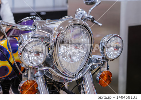 Detailed close-up view of front of custom motorcycle, showcasing chrome components and lighting system. Central headlight is prominent, flanked by smaller lights and striking flame graphic. 140459133