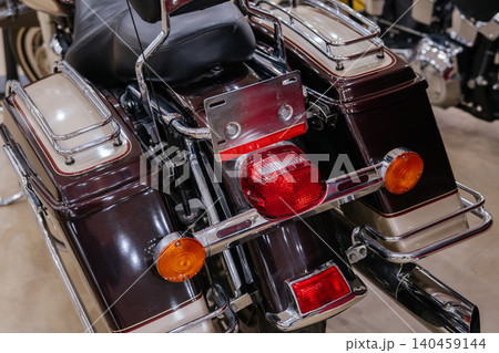 Detailed close-up view of rear section of touring motorcycle, showcasing chrome accents, red taillight, amber turn signals, and burgundy and cream saddlebags. Bright lighting highlights polished 140459144