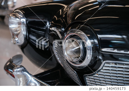 Detailed low-angle view highlights front right fender, headlight, and grille of vintage black automobile. Glossy paint reflects light, showcasing chrome accents and classic design elements. Detailed low-angle view highlights front right fender, headlight, and grille of vintage black automobile. Glossy paint reflects light, showcasing chrome accents and classic design elements. 140459173