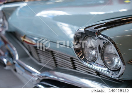 Detailed close-up view of front section of light blue vintage automobile. Polished chrome headlights and grille reflect surroundings, emphasizing classic design in a softly blurred indoor setting. Detailed close-up view of front section of light blue vintage automobile. Polished chrome headlights and grille reflect surroundings, emphasizing classic design in a softly blurred indoor setting. 140459180
