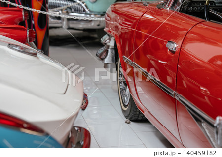 Close-up view of a vintage red car showcasing its shiny chrome details. A white classic car is partially visible in the foreground, while a teal vehicle adds to the nostalgic atmosphere of this indoor Close-up view of a vintage red car showcasing its shiny chrome details. A white classic car is partially visible in the foreground, while a teal vehicle adds to the nostalgic atmosphere of this indoor 140459182
