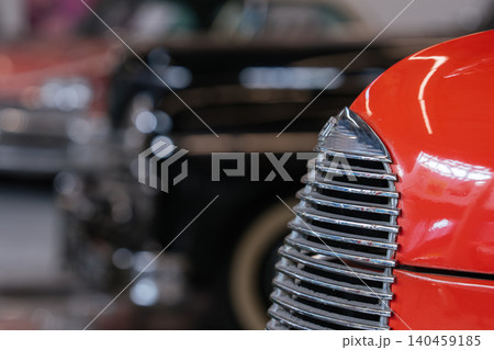 Detailed view highlights polished chrome grille and vibrant orange fender of vintage automobile. Softly blurred background reveals another classic car, enhancing automotive nostalgia. Detailed view highlights polished chrome grille and vibrant orange fender of vintage automobile. Softly blurred background reveals another classic car, enhancing automotive nostalgia. 140459185