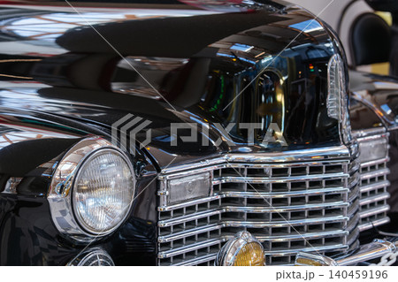 Detailed view of front of classic black car, showcasing polished chrome grille and round headlight. Gleaming exterior reflects indoor exhibition space, highlighting vintage design and craftsmanship. 140459196