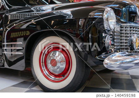Detailed close-up captures vintage black automobile showcasing polished exterior and chrome accents. Front right wheel features whitewall tire with red rim, reflecting indoor setting and checkered 140459198
