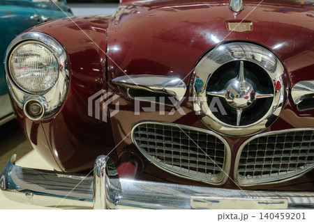 Detailed low-angle view showcases front of classic maroon car. Image highlights chrome grille with starburst emblem, round headlight, and polished chrome bumper, with another vintage car in background Detailed low-angle view showcases front of classic maroon car. Image highlights chrome grille with starburst emblem, round headlight, and polished chrome bumper, with another vintage car in background 140459201
