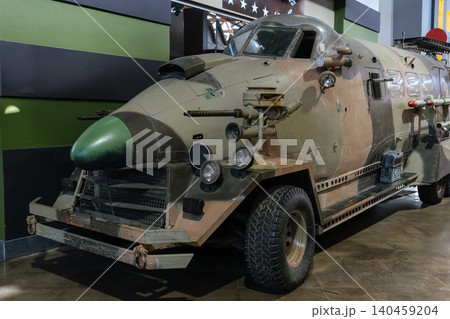 Unique armored military vehicle showcased indoors features distinctive camouflage and multiple mounted weapons. The vehicle includes a green-tipped aerodynamic front, riveted plating, and large off Unique armored military vehicle showcased indoors features distinctive camouflage and multiple mounted weapons. The vehicle includes a green-tipped aerodynamic front, riveted plating, and large off 140459204