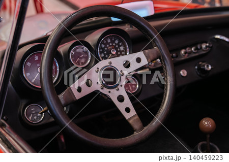 Close-up view of vintage car interior showcasing leather-wrapped steering wheel and circular gauges. Dashboard features black surfaces with metallic accents and wooden gear shift knob, highlighting Close-up view of vintage car interior showcasing leather-wrapped steering wheel and circular gauges. Dashboard features black surfaces with metallic accents and wooden gear shift knob, highlighting 140459223