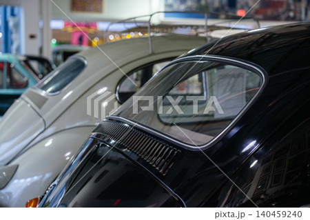 Close-up view of two vintage cars, one glossy black and the other light-colored, parked closely together. The scene highlights their classic design, with reflections of framed pictures visible on the Close-up view of two vintage cars, one glossy black and the other light-colored, parked closely together. The scene highlights their classic design, with reflections of framed pictures visible on the 140459240