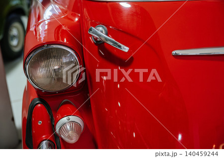 Detailed view of vintage red car's front fender, showcasing glossy paint and chrome features. Highlights include a classic headlight, rounded indicator light, and gleaming door handle. 140459244