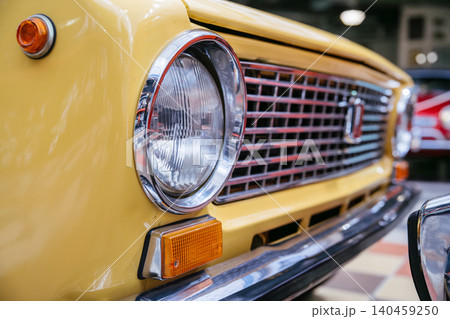 Detailed low-angle view of front of classic yellow automobile, highlighting iconic chrome grille, round headlight with ribbed glass, and amber turn signal. Polished paintwork gleams with soft Detailed low-angle view of front of classic yellow automobile, highlighting iconic chrome grille, round headlight with ribbed glass, and amber turn signal. Polished paintwork gleams with soft 140459250