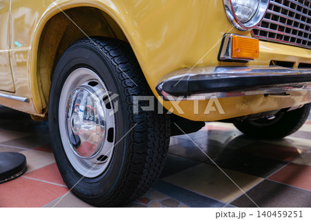 Detailed low-angle view highlights front section of classic yellow automobile. Image showcases textured tire, chrome hubcap, bumper, and grille against a geometric tiled floor. Detailed low-angle view highlights front section of classic yellow automobile. Image showcases textured tire, chrome hubcap, bumper, and grille against a geometric tiled floor. 140459251