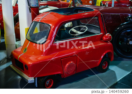 Bright red vintage microcar showcased in museum setting. Rounded design features dark sunroof, chrome accents, and white steering wheel, surrounded by classic vehicles and a fire extinguisher. Bright red vintage microcar showcased in museum setting. Rounded design features dark sunroof, chrome accents, and white steering wheel, surrounded by classic vehicles and a fire extinguisher. 140459260
