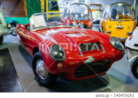 Vintage red convertible sports car prominently displayed indoors, likely in a museum or exhibition. Classic design features rounded headlights and a distinctive grille, with colorful microcars in the Vintage red convertible sports car prominently displayed indoors, likely in a museum or exhibition. Classic design features rounded headlights and a distinctive grille, with colorful microcars in the 140459261