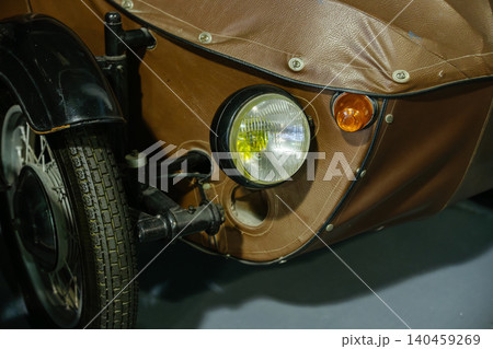 Detailed close-up view of vintage motorcycle sidecar showcasing brown textured covering and chrome headlight. Small orange indicator light and part of front wheel visible, emphasizing classic design. 140459269