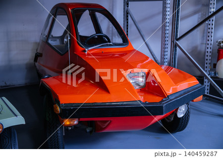 Vintage three-wheeled microcar in bright orange color is parked indoors, showcasing its unique bubble canopy and minimalist design. Industrial shelving in the background adds to the retro charm of Vintage three-wheeled microcar in bright orange color is parked indoors, showcasing its unique bubble canopy and minimalist design. Industrial shelving in the background adds to the retro charm of 140459287