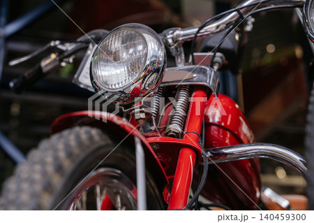Detailed low-angle view of front of vintage red motorcycle. Focus on chrome headlight, handlebars, and suspension springs, evoking nostalgia for classic machinery in a blurred indoor setting. Detailed low-angle view of front of vintage red motorcycle. Focus on chrome headlight, handlebars, and suspension springs, evoking nostalgia for classic machinery in a blurred indoor setting. 140459300