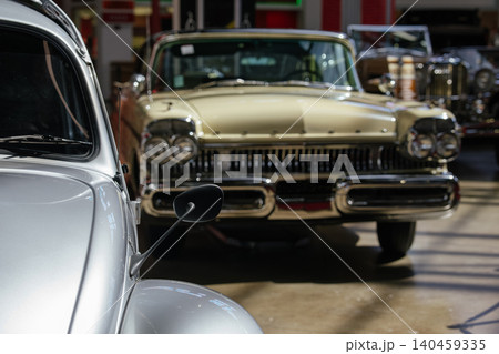 Vintage cream-colored sedan stands prominently in classic car showroom. Silver compact car partially visible in foreground, evoking nostalgia and polished elegance of bygone eras. 140459335