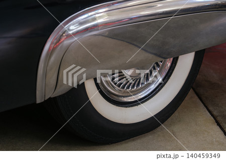 Detailed view captures rear wheel and fender of classic car. Distinct white sidewall tire and polished chrome trim highlight vintage automotive craftsmanship against textured ground. Detailed view captures rear wheel and fender of classic car. Distinct white sidewall tire and polished chrome trim highlight vintage automotive craftsmanship against textured ground. 140459349
