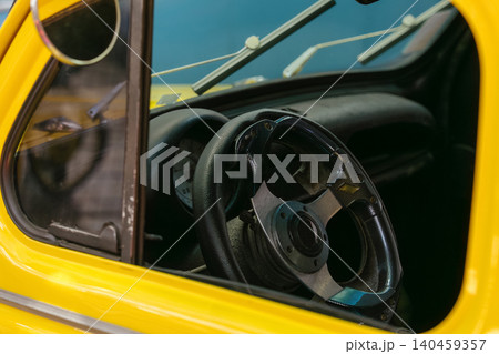 Detailed close-up of vintage yellow car interior viewed through side window. Worn aftermarket steering wheel features metallic hub and textured grip, evoking nostalgia and automotive history. Detailed close-up of vintage yellow car interior viewed through side window. Worn aftermarket steering wheel features metallic hub and textured grip, evoking nostalgia and automotive history. 140459357