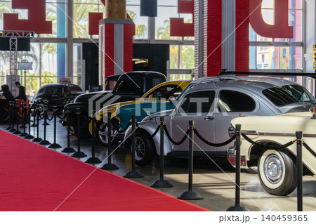 Vintage car exhibition showcases classic vehicles on a polished floor. A vibrant red carpet leads towards a lineup of antique cars, with palm trees visible through large windows. Vintage car exhibition showcases classic vehicles on a polished floor. A vibrant red carpet leads towards a lineup of antique cars, with palm trees visible through large windows. 140459365