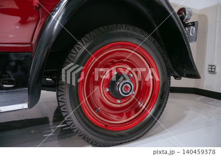 Detailed close-up captures vintage automobile wheel featuring glossy red rim and classic black tire. Low-angle perspective highlights design elements, polished floor, and visible license plate. 140459378