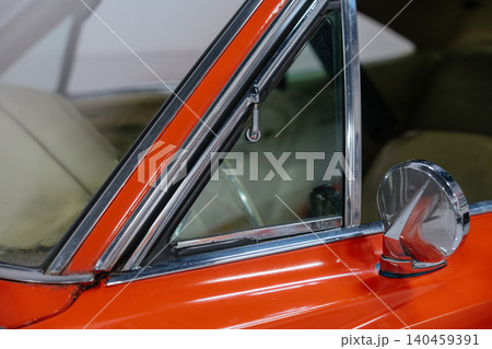 Close-up view highlights vintage car's triangular vent window with vibrant orange paint and polished chrome accents. Round side mirror attached to door reflects ambient details, showcasing classic Close-up view highlights vintage car's triangular vent window with vibrant orange paint and polished chrome accents. Round side mirror attached to door reflects ambient details, showcasing classic 140459391