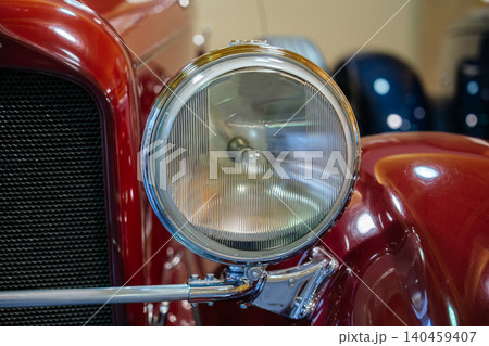 Detailed close-up view of classic red car's headlight and grill. Round headlight features ribbed glass lens and chrome trim, while polished red paint reflects surrounding light. Detailed close-up view of classic red car's headlight and grill. Round headlight features ribbed glass lens and chrome trim, while polished red paint reflects surrounding light. 140459407