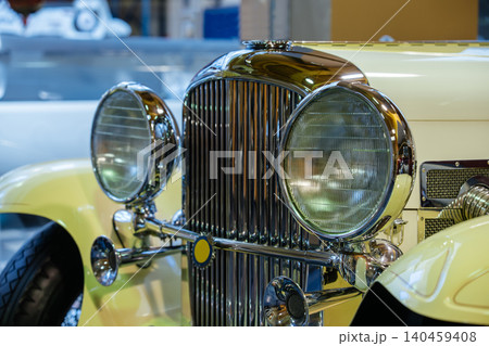 Detailed close-up view of front of vintage yellow car. Polished chrome grille and large round headlights with ribbed glass create a striking automotive design in an exhibition setting. Detailed close-up view of front of vintage yellow car. Polished chrome grille and large round headlights with ribbed glass create a striking automotive design in an exhibition setting. 140459408