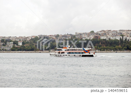 A cruise boat glides across the Bosphorus, surrounded by lush hills and modern buildings, under a cloudy sky that hints at possible rain. A cruise boat glides across the Bosphorus, surrounded by lush hills and modern buildings, under a cloudy sky that hints at possible rain. 140459651