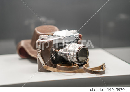 A vintage camera sits on display in a museum, showcasing its unique design with a leather strap and case, celebrating photographys past. 140459704