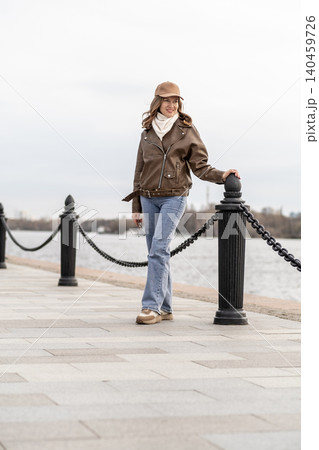 A woman walks along a waterfront promenade, wearing a brown leather jacket and cap, with a relaxed expression against a cloudy sky. 140459726
